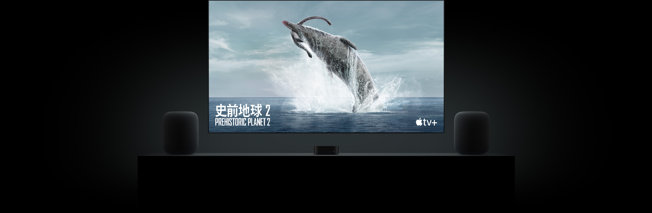 螢幕寬闊的平面電視顯示節目《史前地球》中鮮明逼真的恐龍畫面。電視掛牆,下方的客廳几櫃上有一部 Apple TV,兩旁各有一部 HomePod 揚聲器
